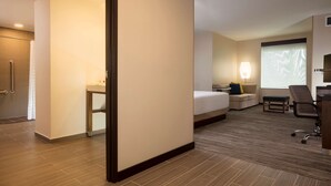 Quarto de hotel com cama, escrivaninha, cadeira, luminária e janela com persianas.
