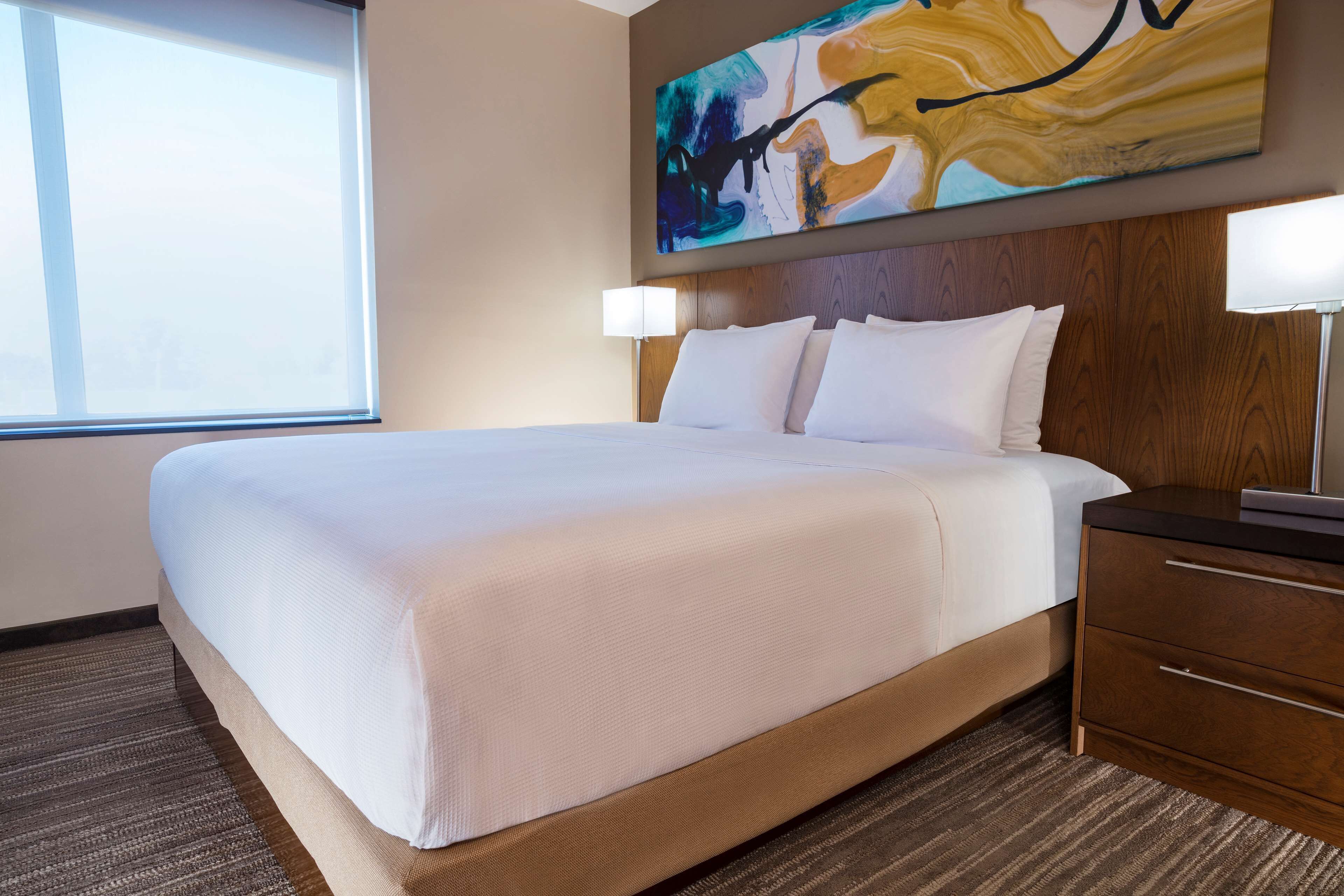 Habitación de hotel con una cama grande, cabecera de madera, mesita de noche con lámpara y un cuadro sobre la cama.
