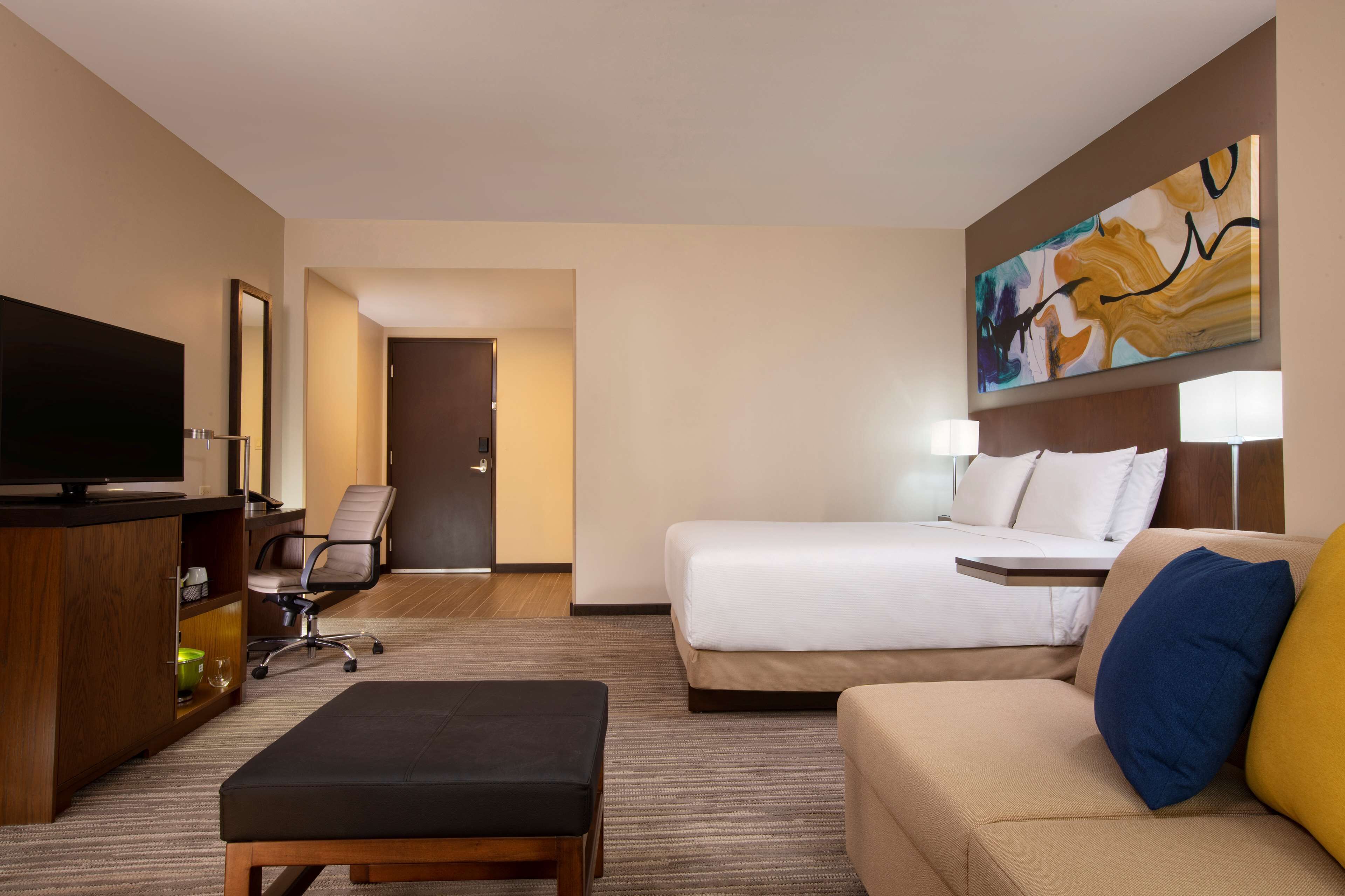 A hotel room with a bed, a sofa, a TV, a desk, and a painting on the wall.