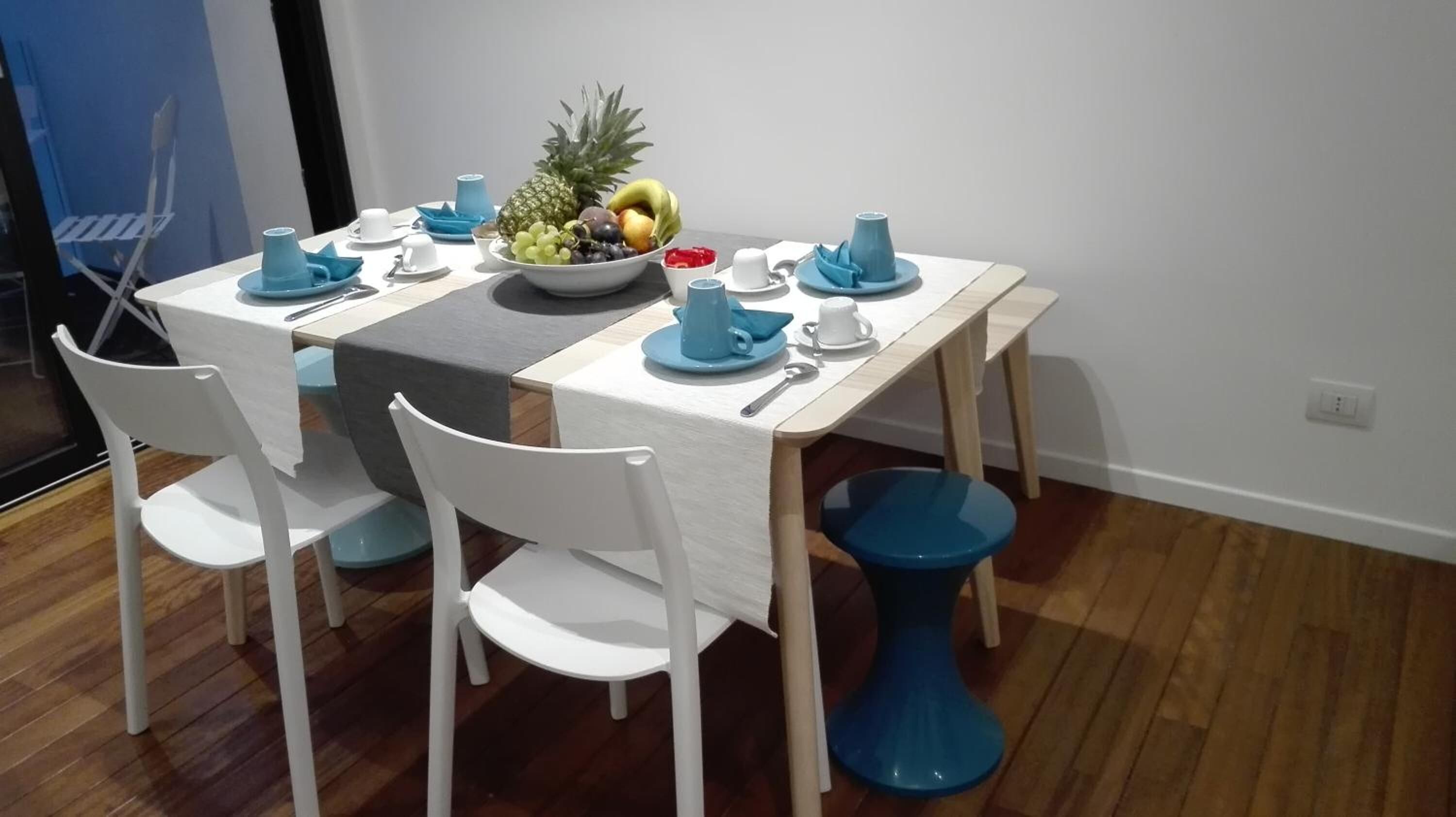 A dining area with a table set for two, featuring white chairs, blue tableware, and a fruit bowl.
