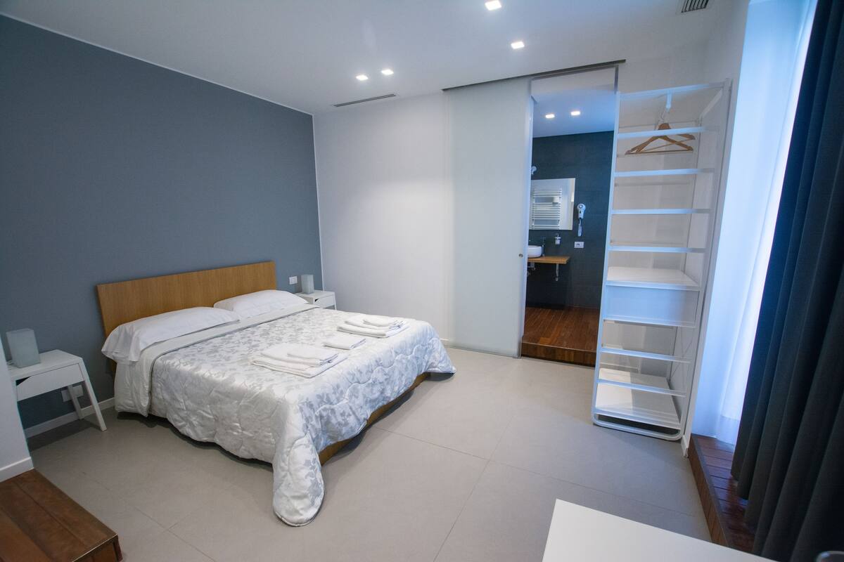 Une chambre à coucher moderne, avec un lit, des tables de chevet, un dressing et une salle de bain visible à travers une porte ouverte.