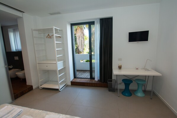 A modern hotel room with a walk-in closet, a small desk with two stools, a television, and a sliding glass door to the balcony.