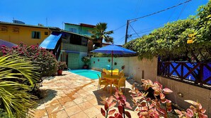 A backyard with a swimming pool, a patio table with chairs, and a blue umbrella. - Pousada Paranambuca (Ipojuca)