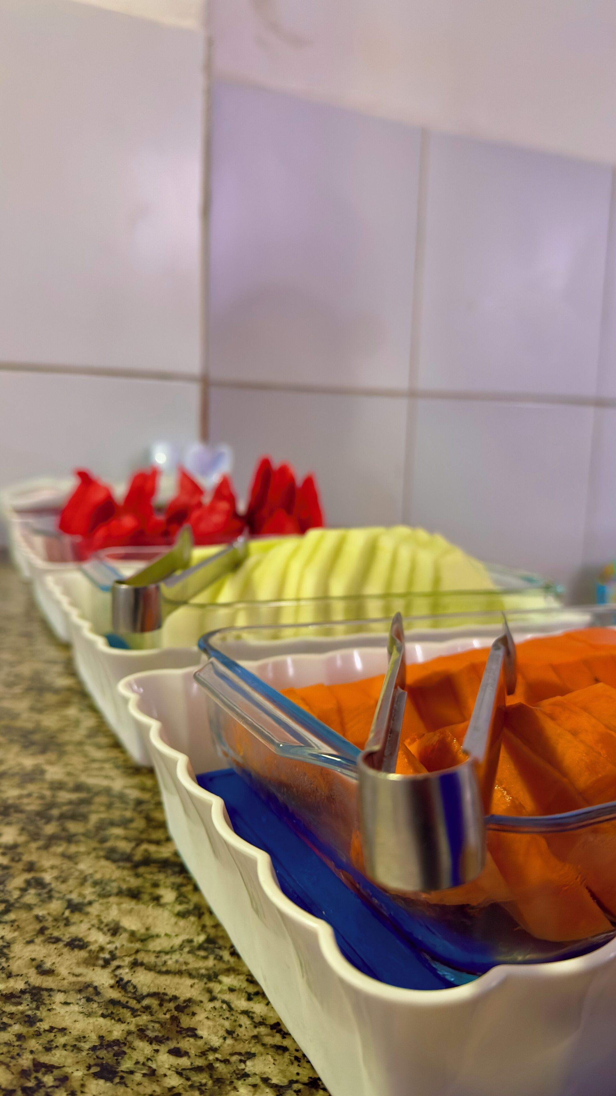 Buffet com diversos alimentos em recipientes transparentes, incluindo frutas e uma sobremesa amarela.