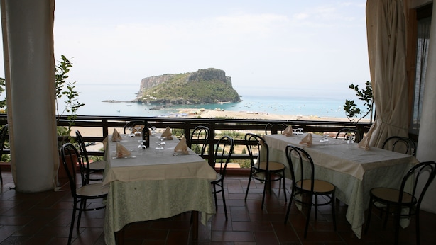 An outdoor dining area with tables and chairs, overlooking a beach and a rocky island. - Hotel i Normanni (Praia a Mare)