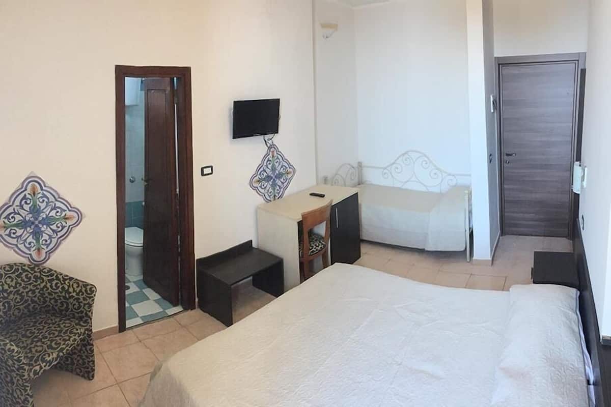 A hotel room with a bed, a desk, a chair, a TV, and a bathroom visible through an open door.