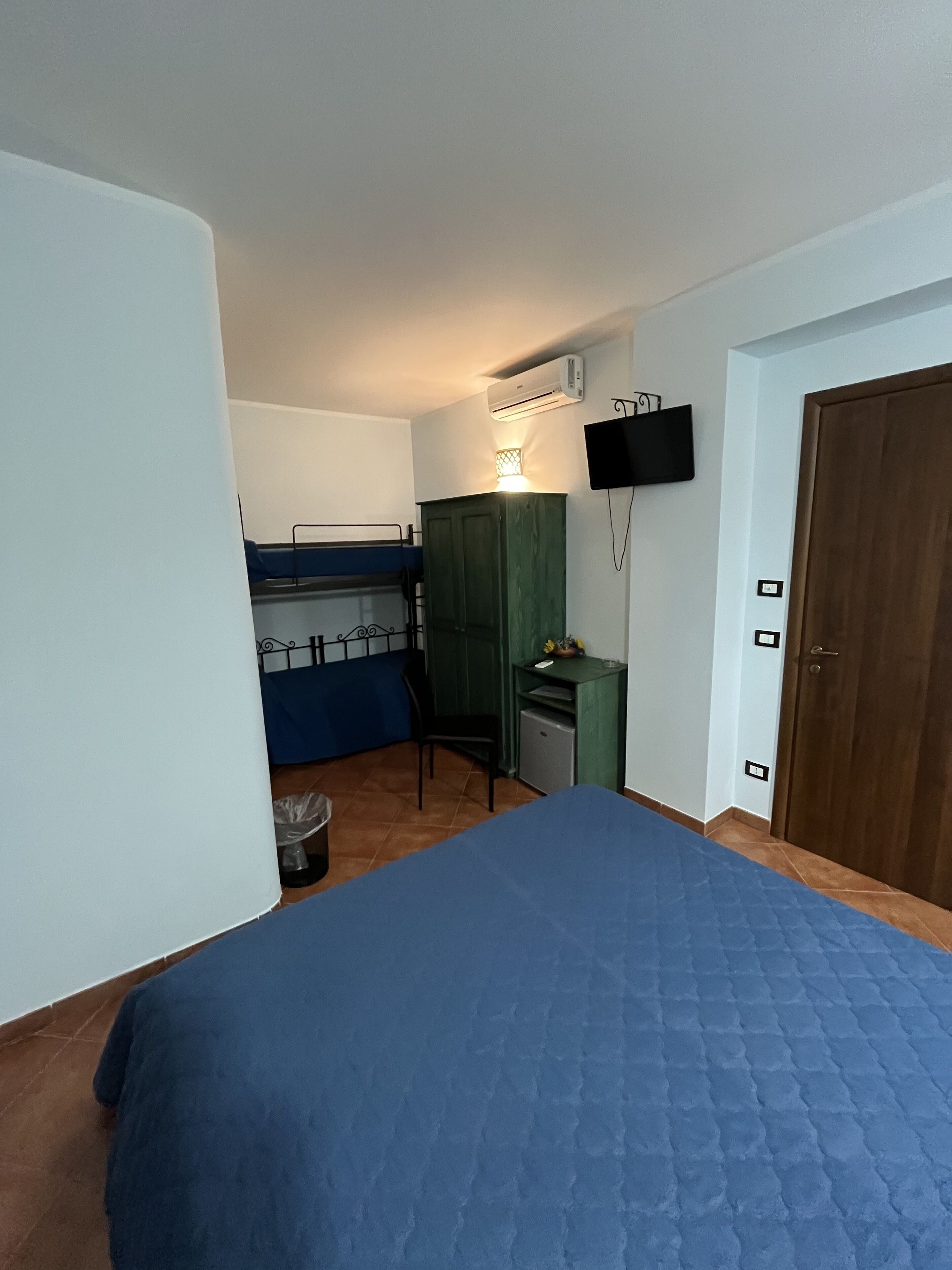 A compact room with a bunk bed, a desk, a chair, a small cabinet, a television mounted on the wall, and a door.