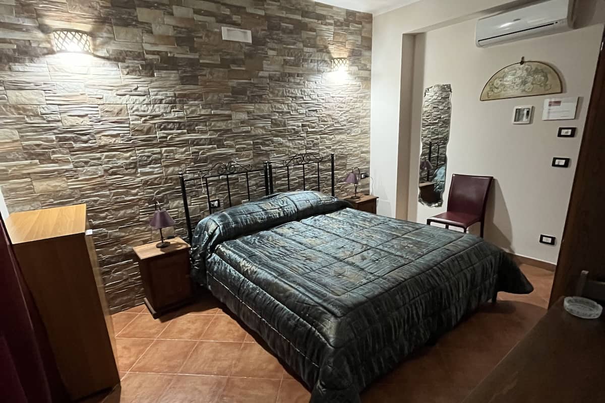 Un dormitorio con pared de diseño de piedra, una cama con colcha oscura, una mesita de noche, una lámpara, una silla y una mesa de madera.