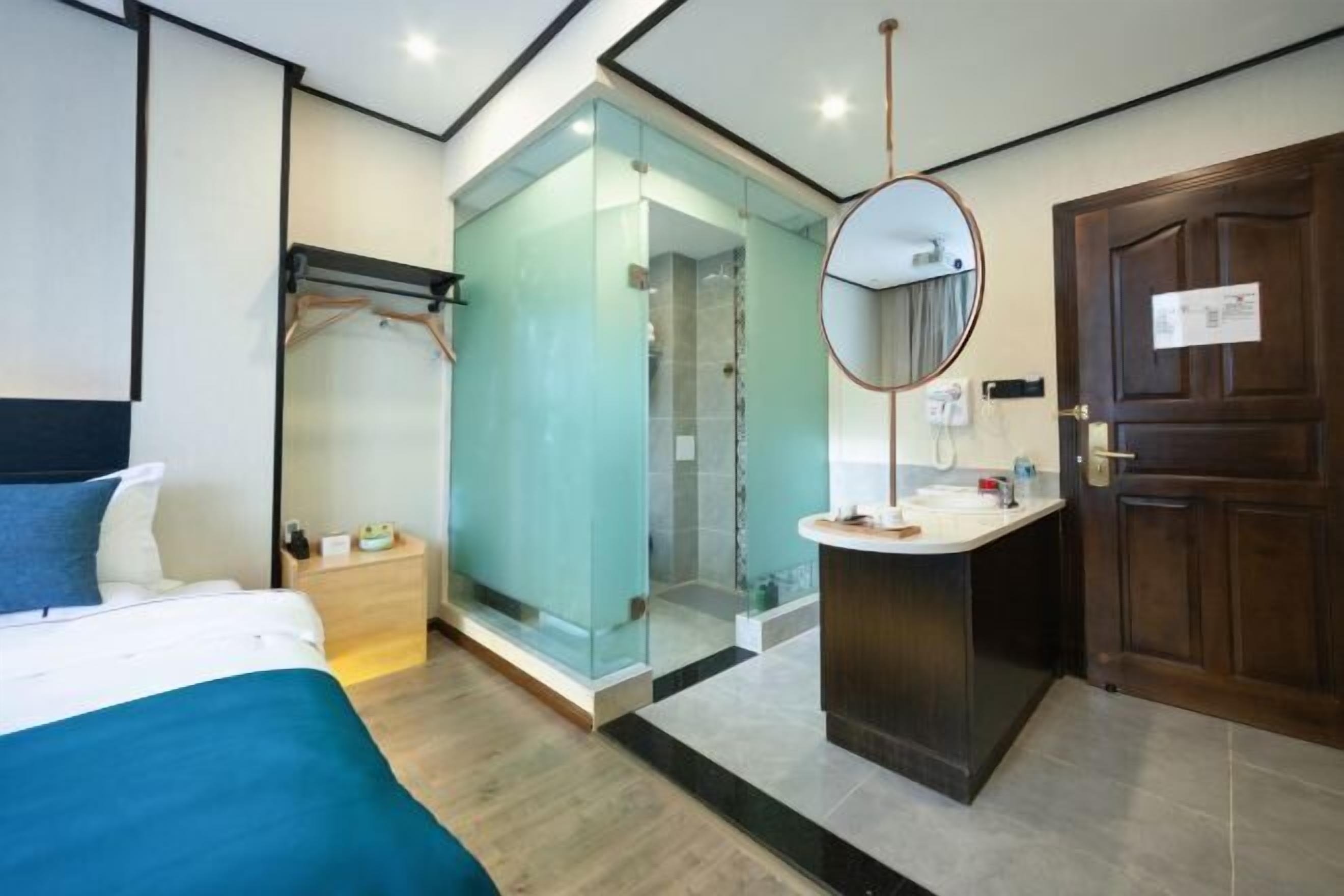 Una habitación de hotel moderna con una cama, una zona de ducha con paredes de vidrio, un espejo redondo y una puerta de madera.