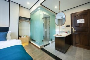 A modern hotel room with a bed, a glass-enclosed shower area, a round mirror, and a wooden door.