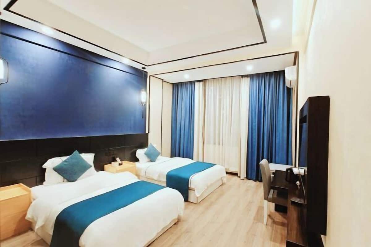 A hotel room with two beds, a desk, a chair, a TV, and a large window with curtains.