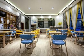 A modern dining area with wooden tables and blue chairs, a kitchen in the background, and a view of the city through large windows. - Hongsen Business Hotel (Tengchong)