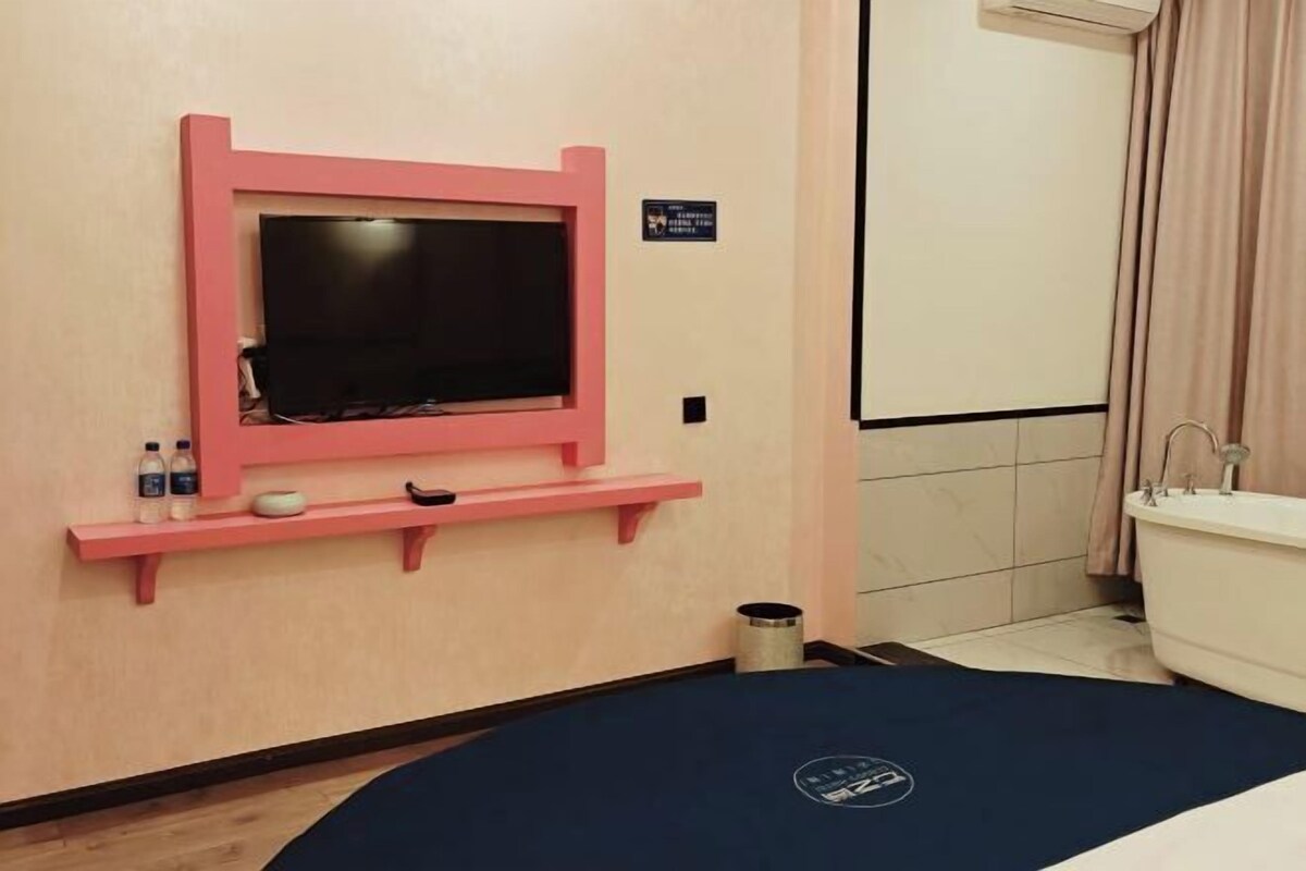 A hotel room with a bed, a TV mounted on a pink shelf, a small trash bin, and a bathroom with a bathtub and a towel rack.