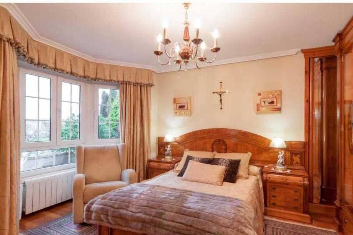 Un dormitorio con una cama grande, una silla, una mesita de noche de madera, una araña y una ventana con cortinas.