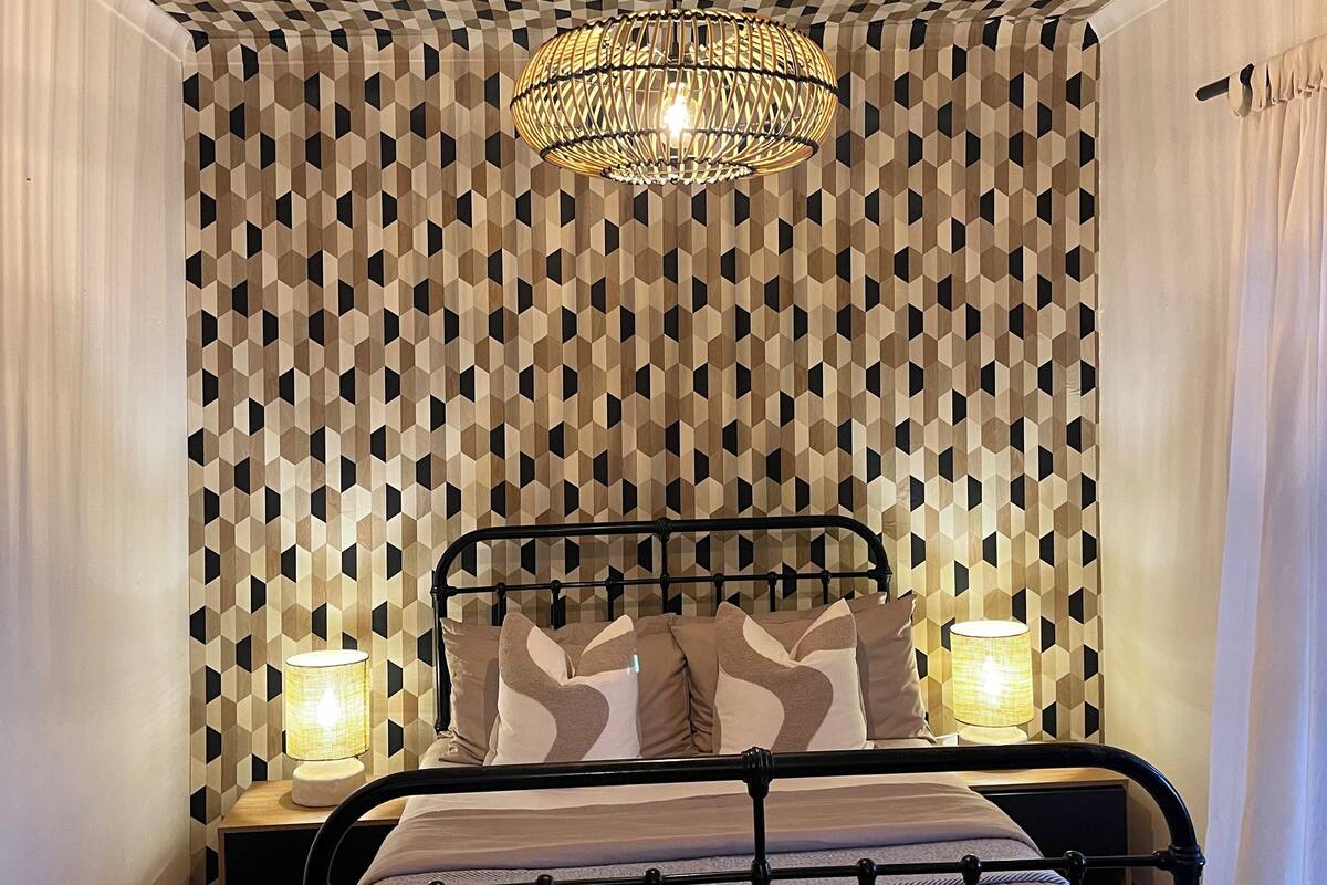 A bedroom with a geometric patterned ceiling, a bed with a metal frame, two bedside lamps, and a hanging light fixture.