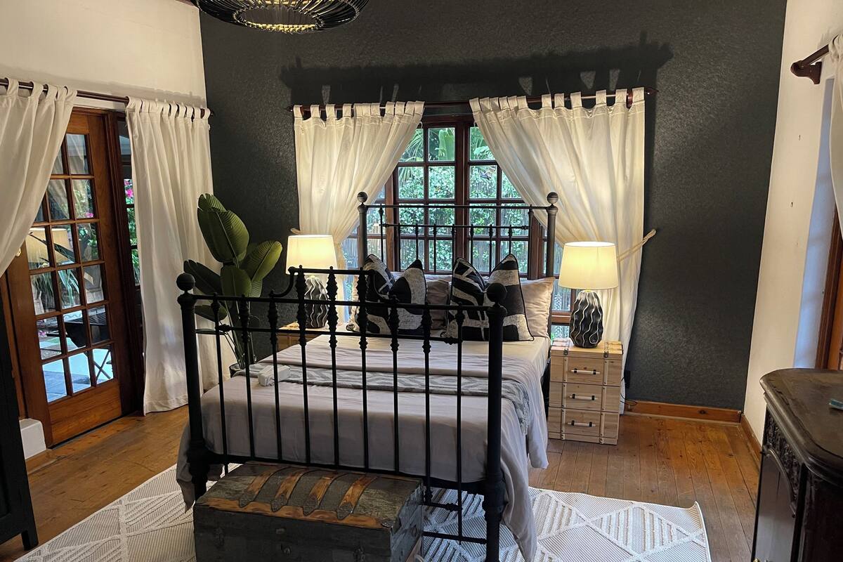 A bedroom with a four-poster bed, two bedside tables with lamps, a wooden chest, and a view through the window.