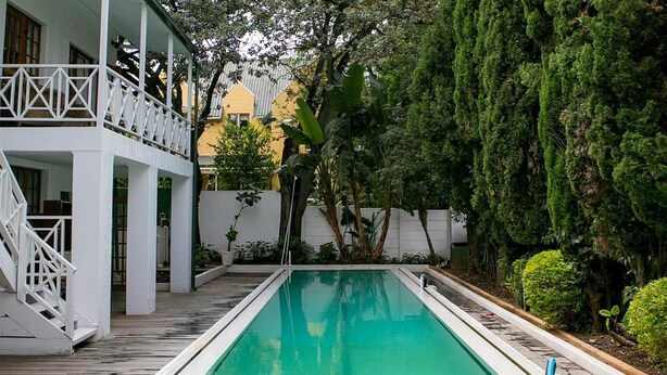 Ein rechteckiger Swimmingpool mit einer Holzterrasse darum herum, ein weißer Balkon mit Geländer und üppiges Grün im Hintergrund.