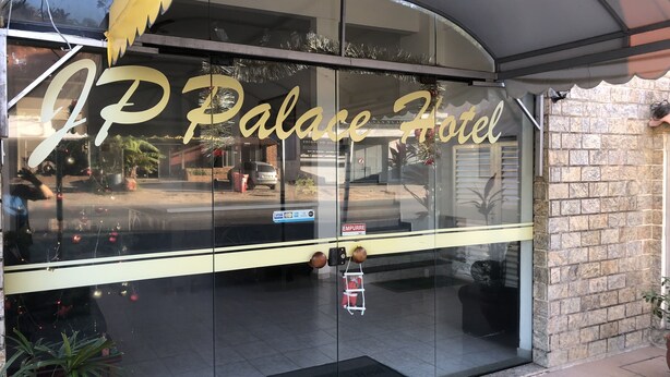 Exterior do Hotel JP Palace, com entrada de vidro, área de estacionamento visível e um muro de tijolos à direita.