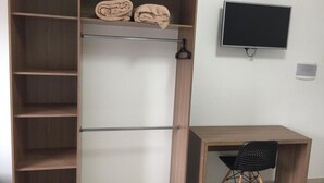Quarto com escrivaninha de madeira, cadeira preta, televisão na parede e nichos embutidos com toalhas dobradas.