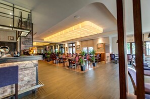 Un amplio vestíbulo de hotel con techo alto, una recepción, varias zonas de comedor con mesas y sillas, y plantas en macetas.