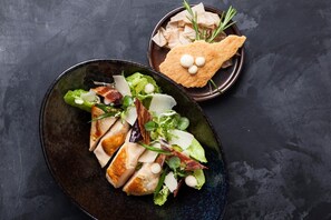 Un plato de pechuga de pollo en rodajas con lechuga y queso, acompañado de un tazón pequeño de crotones y una ramita de romero.