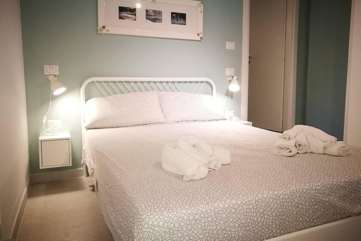 A neatly made bed with a patterned bedspread, two bedside lamps, a small bedside table with a lamp, and a framed picture above the bed.