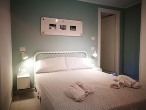 Cama bem arrumada com cabeceira branca, mesa de cabeceira com abajur e um quadro na parede.