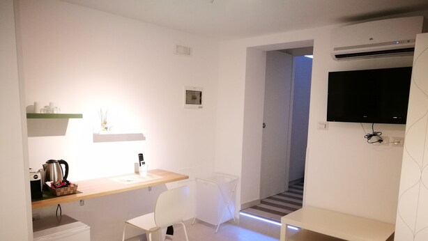 A hotel room with a kitchenette, a desk, a chair, a white bathrobe on the floor, and a TV mounted on the wall. - Simply House in Bari 2 (Bari)