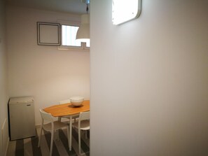 Quarto pequeno e compacto com uma mesa de jantar de madeira e quatro cadeiras brancas. Uma geladeira branca pequena está encostada na parede, e há uma janela acima da mesa.
