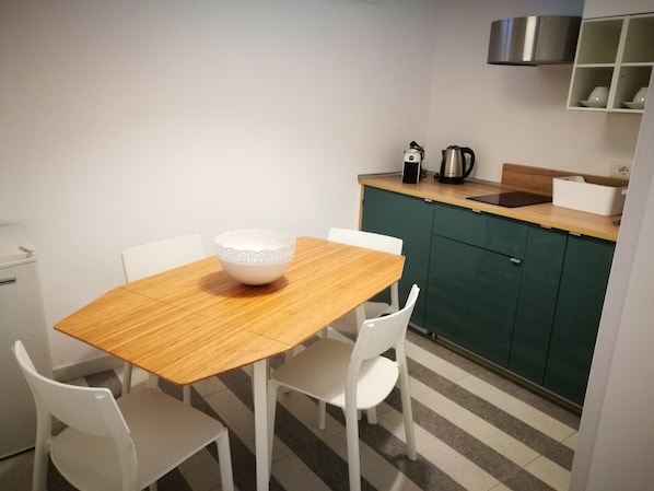 Uma cozinha pequena com uma mesa de madeira e cadeiras brancas, uma chaleira e uma cafeteira no balcão.