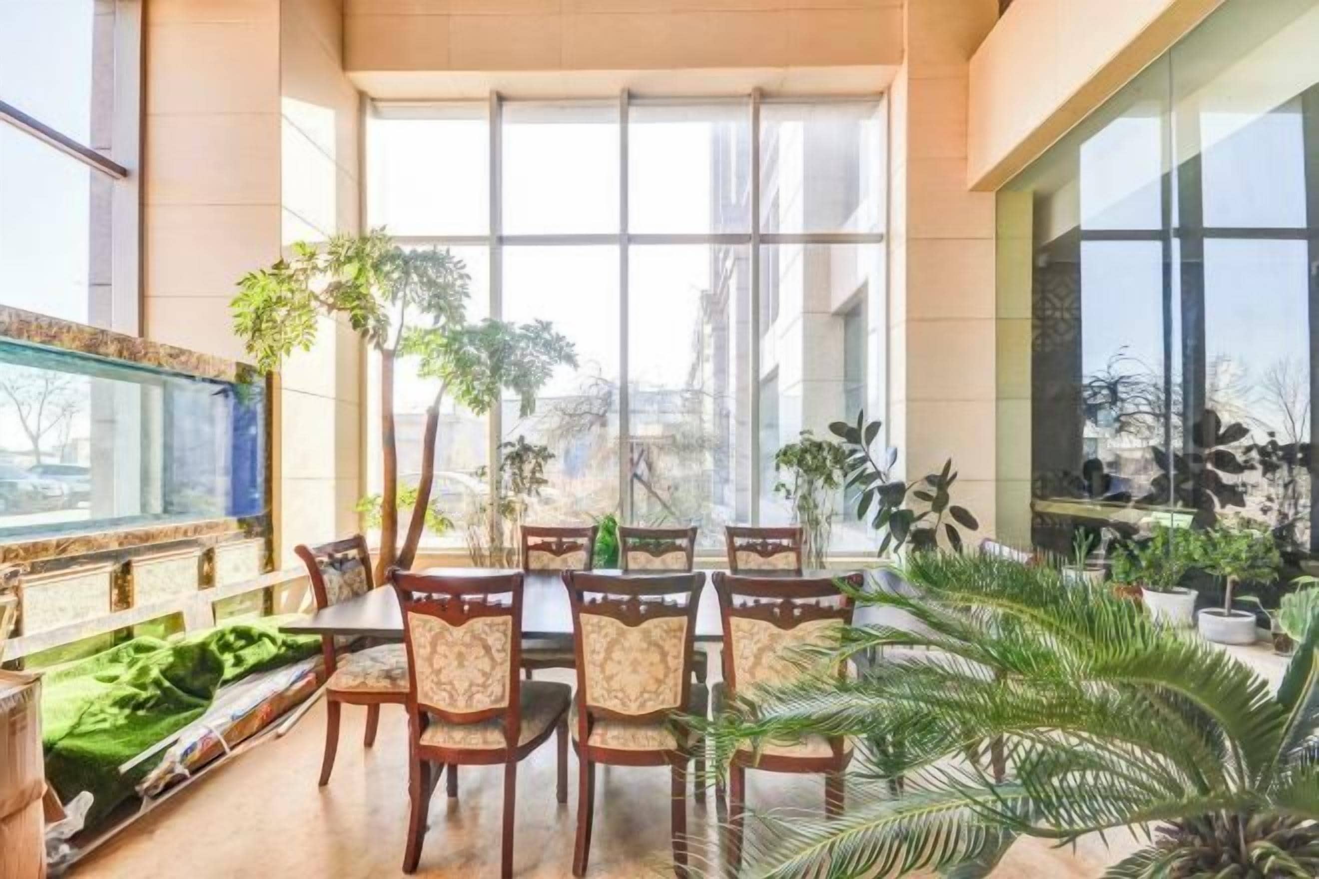 A spacious dining area with a long table and chairs, surrounded by potted plants and large windows.