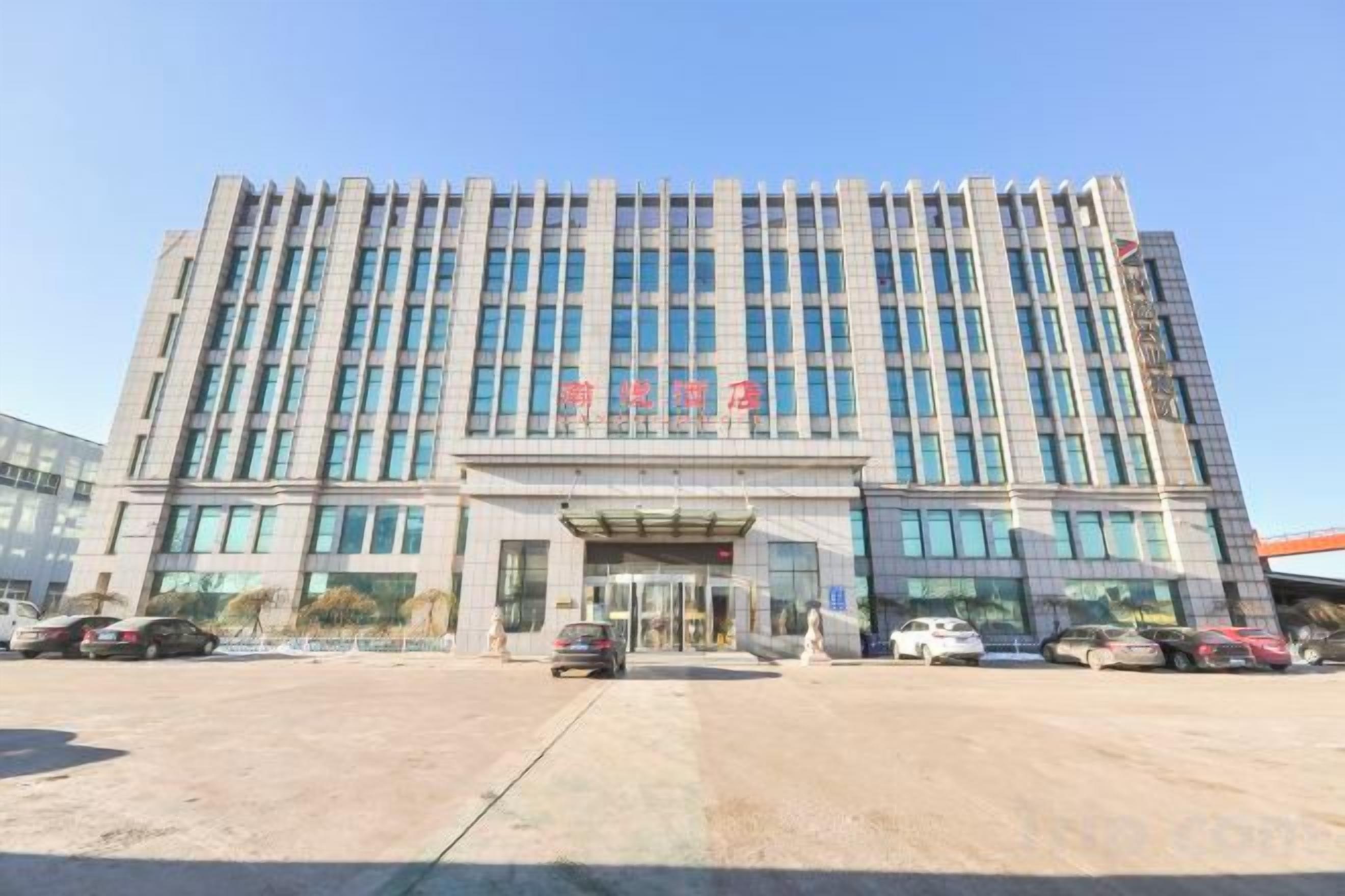 Un edificio de varios pisos con entrada central, ventanas amplias y un estacionamiento con varios autos.