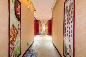 A corridor with patterned carpet, framed artwork on the walls, and a sign indicating the direction to the elevator. - Han Yue Hotel (Tangshan)