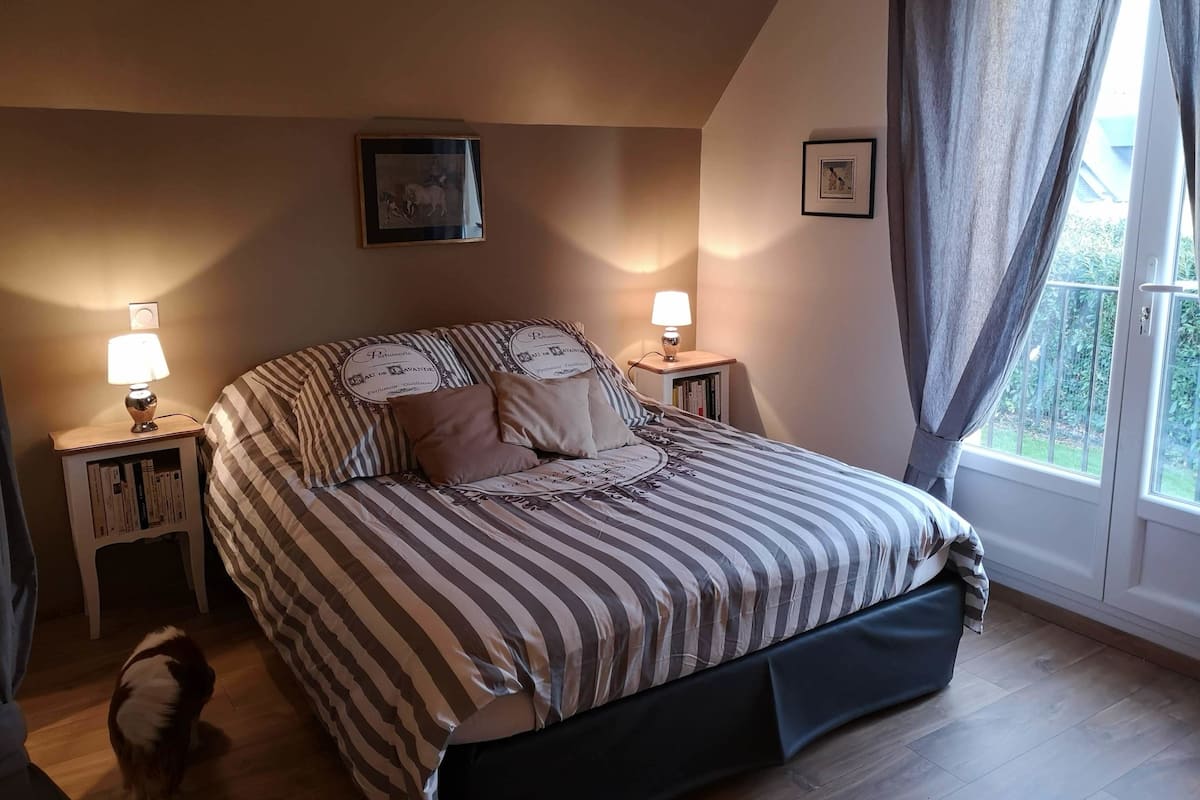 Een slaapkamer met een bed, twee nachtkastjes met lampen, een raam met gordijnen en een foto in een lijst aan de muur.