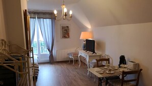 A cozy room with a dining table set for two, a white desk with a lamp, a television, and a radiator. A staircase with a metal railing leads to another room.