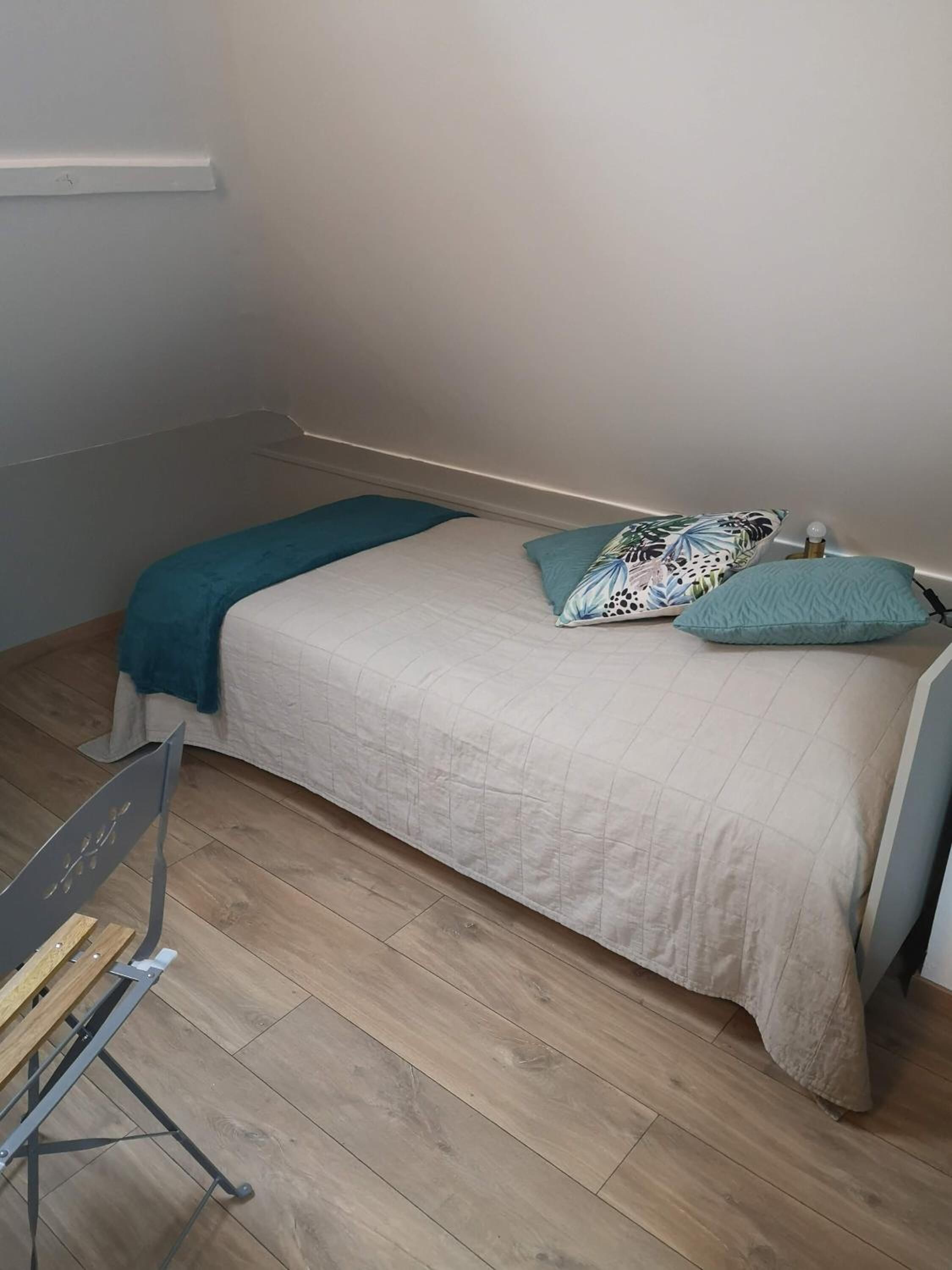 Un petit lit bien fait, avec une couvre-lit beige et deux oreillers : l’un orné d’un motif de feuilles, l’autre d’une couleur bleu-vert unie. La pièce est dotée d’un parquet en bois clair et présente un aménagement simple et fonctionnel.