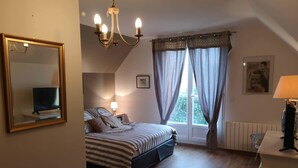 A bedroom with a bed, a bedside table with a lamp, a chair, a table with a cup, a mirror, a chandelier, and a window with curtains.