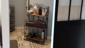 A bathroom with a tiled floor, a metal shelf with towels, toiletries, and a white towel rack.