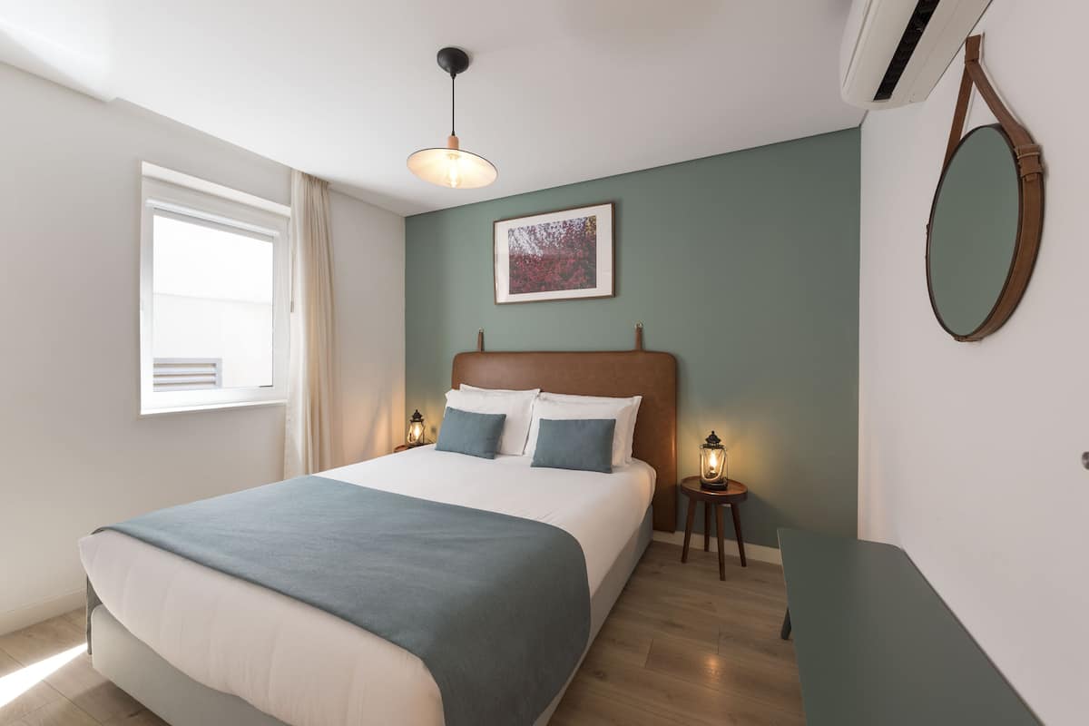 A bedroom with a bed, bedside lamps, a wall-mounted mirror, and a window with curtains.