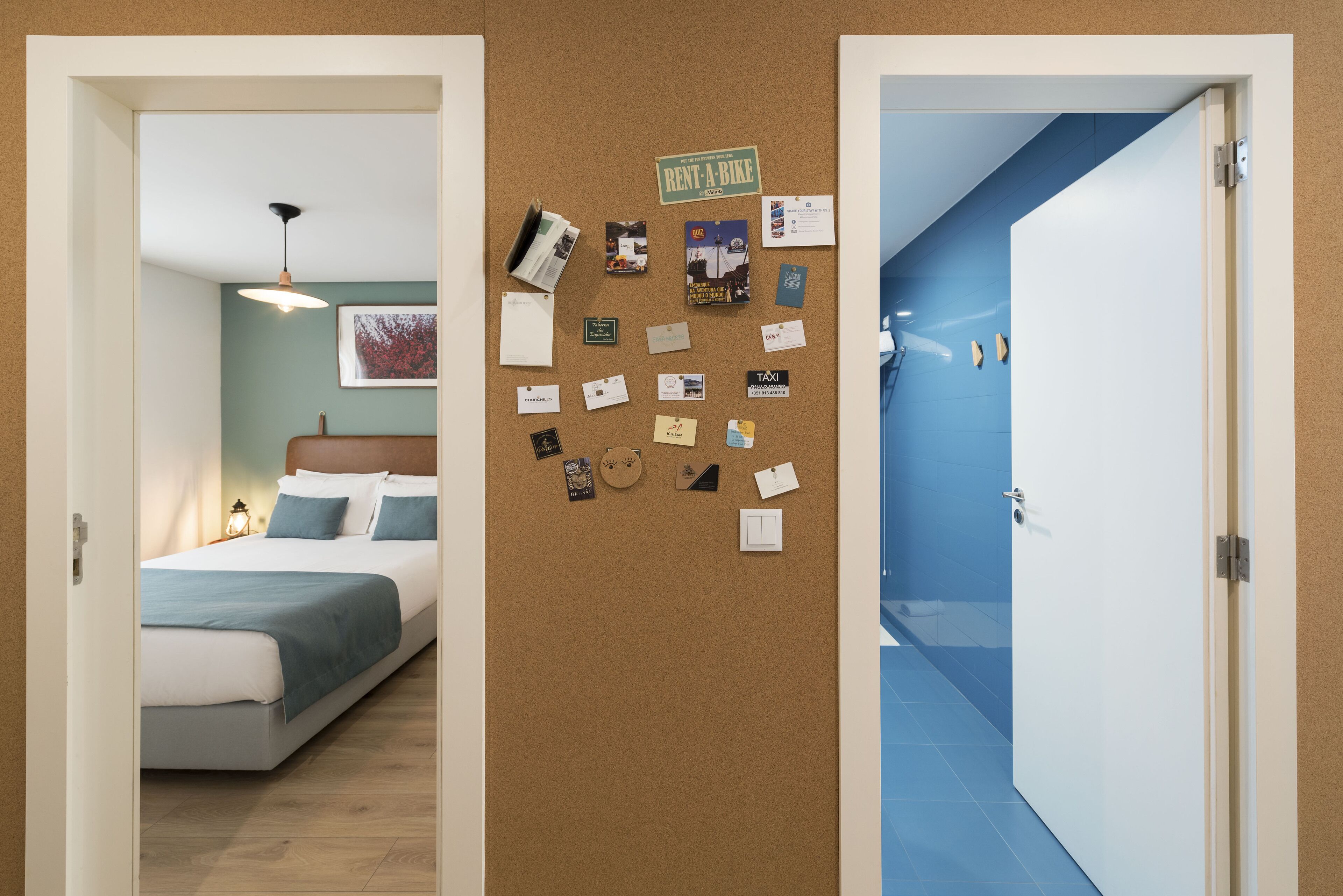 Une chambre d’hôtel avec un lit, un panneau d’affichage mural en liège où sont accrochés divers objets, et une salle de bain avec un mur bleu et une porte blanche.