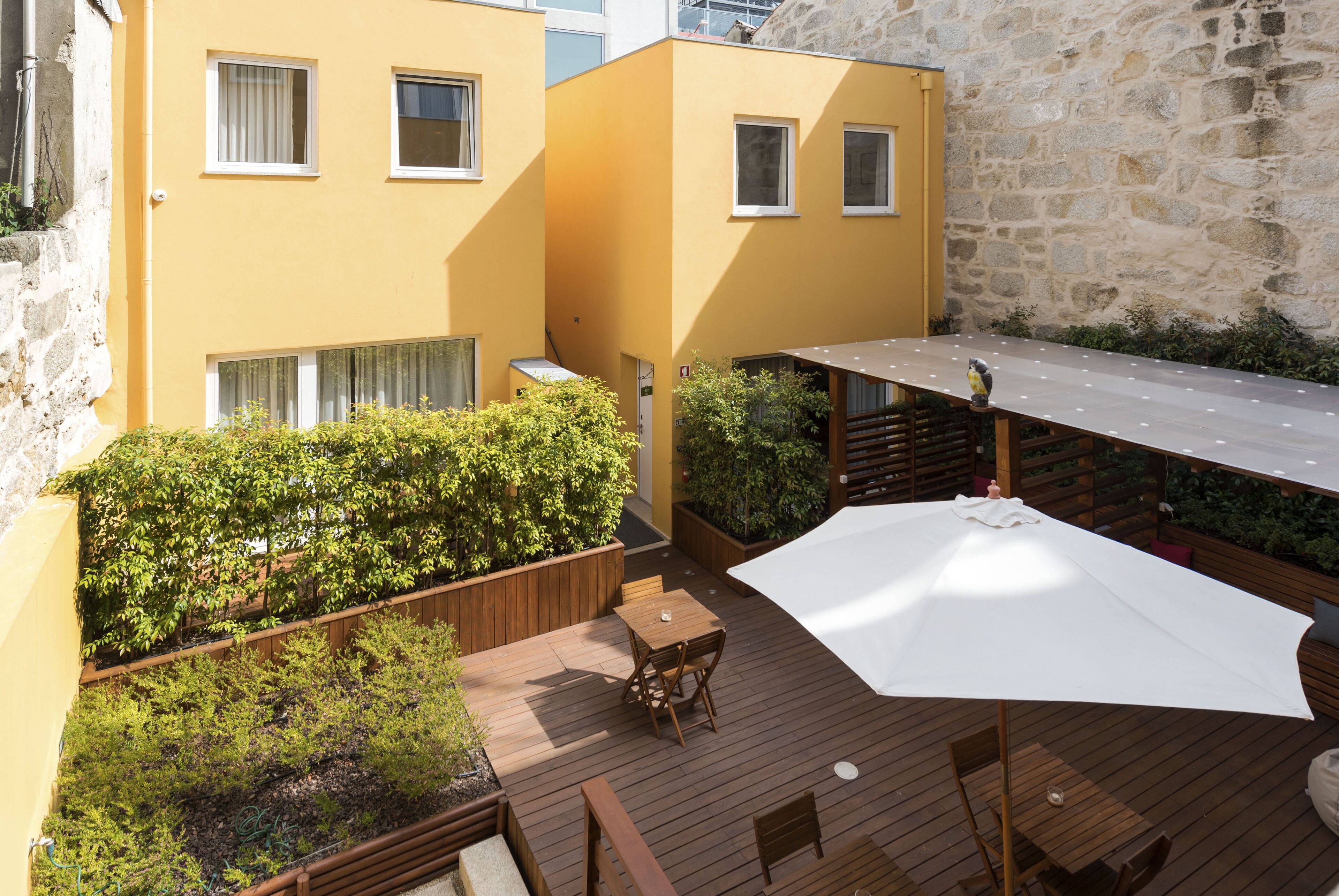 A wooden deck with a table and chairs, surrounded by greenery and a stone wall.