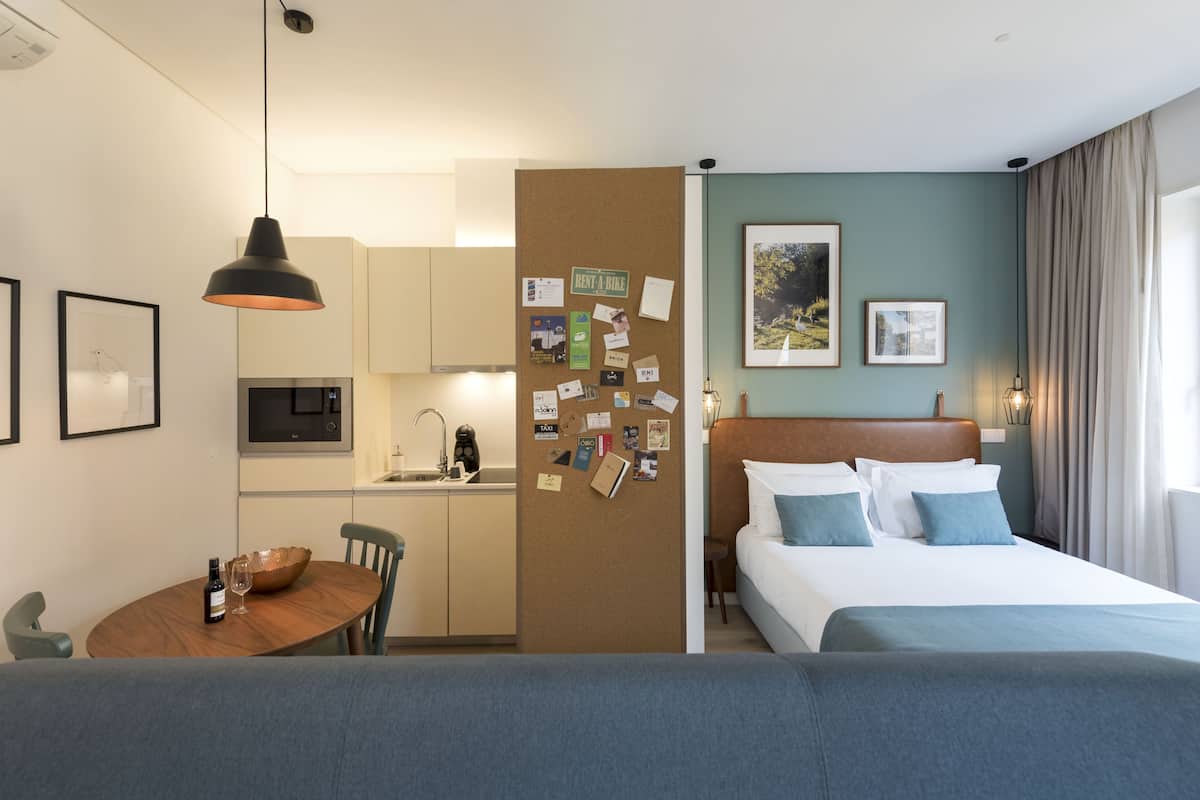 A hotel room with a bed, a desk with a bowl, a corkboard with notes, a kitchen area, and a dining area.