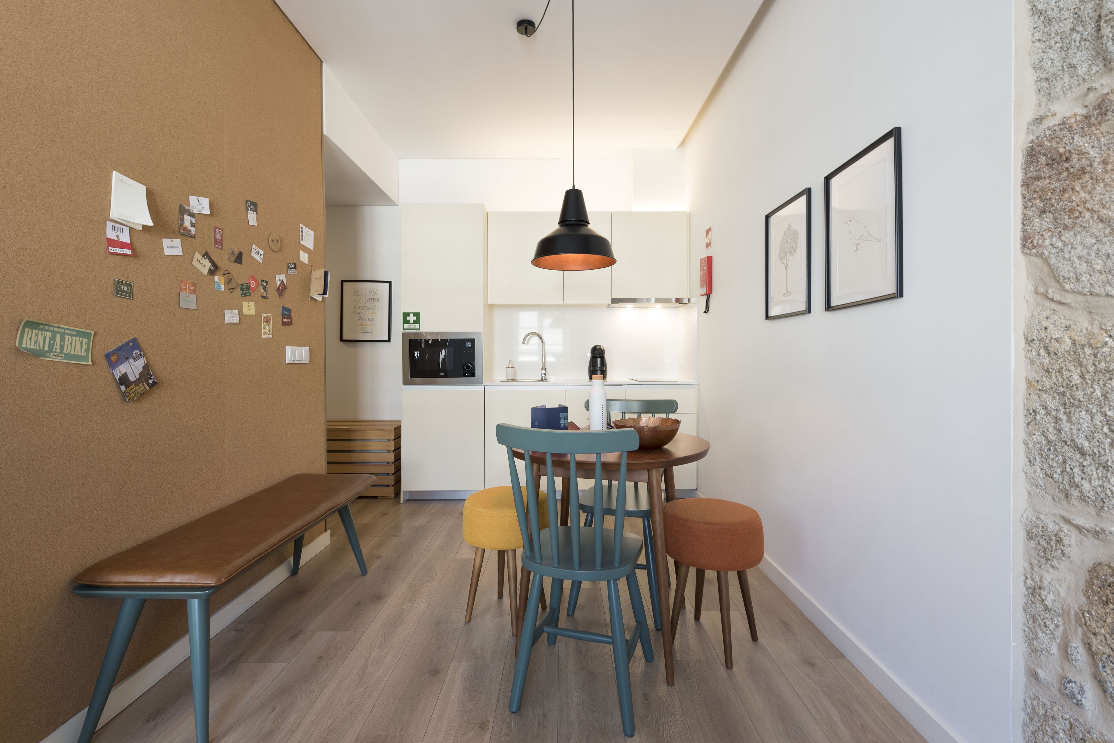 Un intérieur chaleureux avec un banc en bois, une table à manger avec des chaises, un coin cuisine et un tableau d’affichage en liège où sont accrochées des notes.