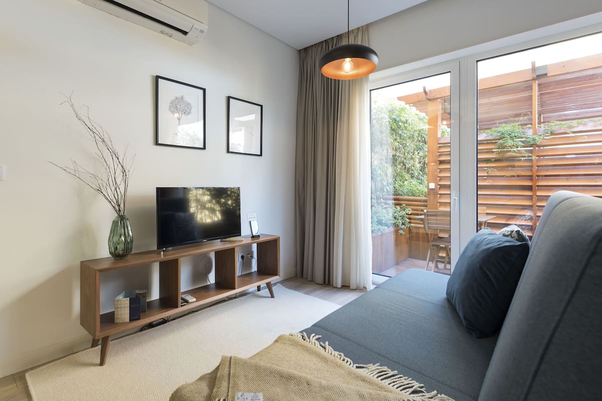 A modern living room with a sofa, a TV on a wooden console, a large window with curtains, and a view of a wooden deck.