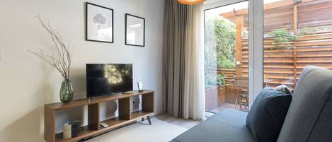 Un salón moderno con un sofá, un televisor sobre una consola de madera, un ventanal con cortinas y vistas a una terraza de madera.