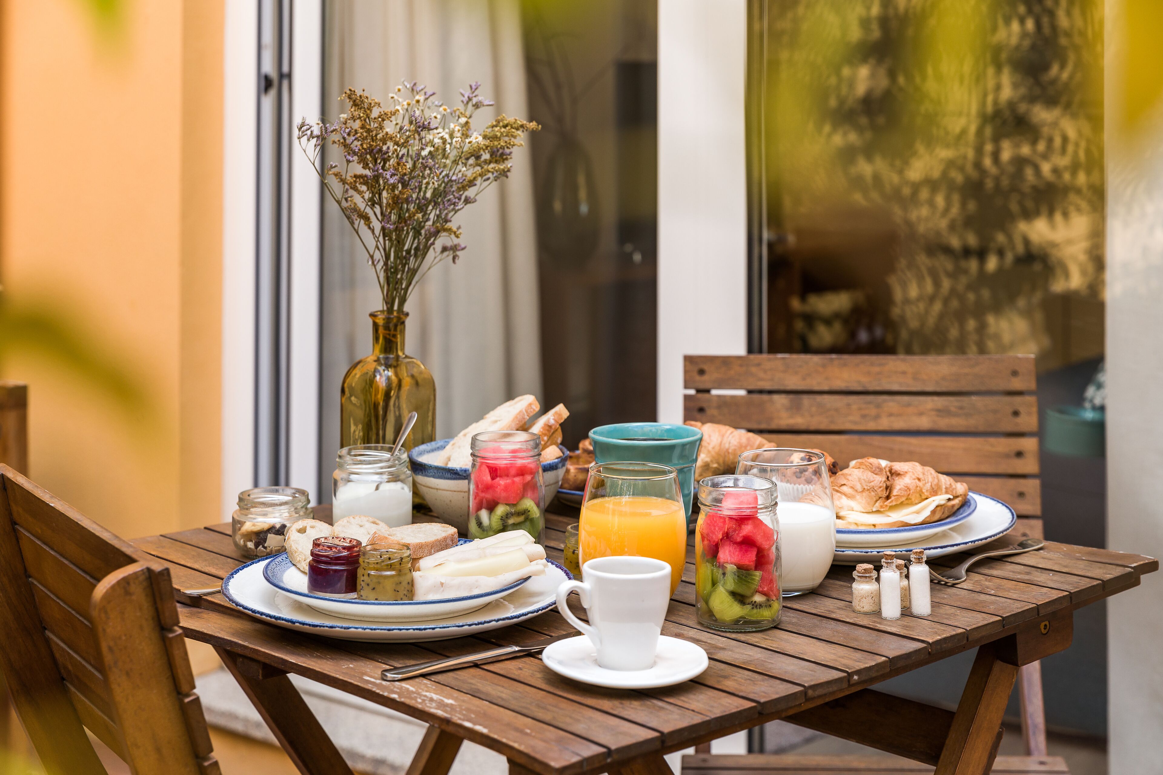 Et træbord dækket med morgenmadsprodukter, blandt andet brød, frugt og drikkevarer, med en vase blomster og saltkar på en solrig terrasse.