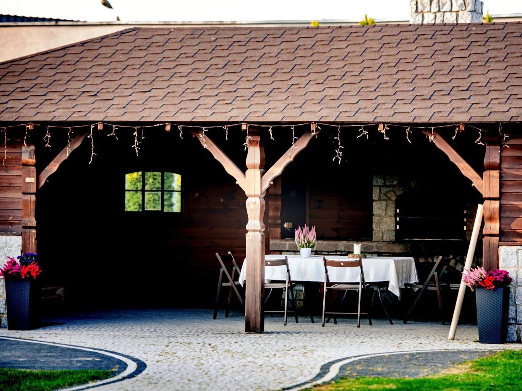 A rustic outdoor dining area with wooden tables and chairs, a stone wall, and a covered entrance.