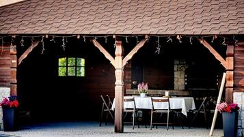 A rustic outdoor dining area with wooden tables and chairs, a stone wall, and a covered entrance.
