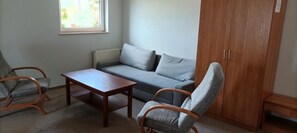 Quarto com sofá cinzento, duas cadeiras de madeira, uma pequena mesa de madeira e um guarda-roupa.