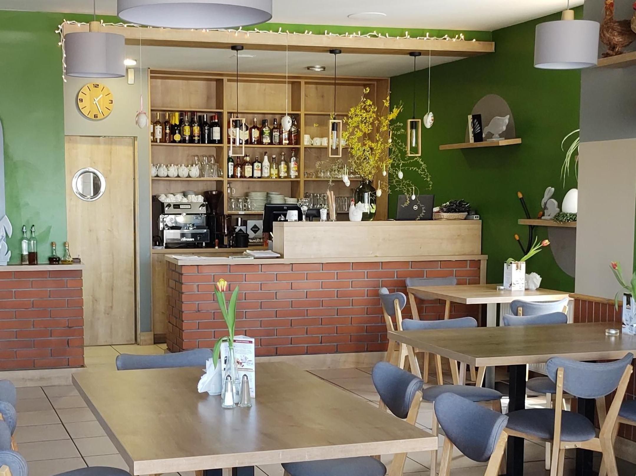 A restaurant interior with wooden tables, blue chairs, a bar area with shelves of bottles, and a green accent wall.